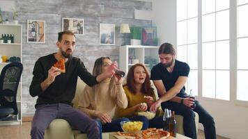 groep van vrienden vieren zege van Amerikaans voetbal bij elkaar passen in leven kamer. jong Mens juichen en Holding een pizza. video