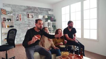 Young man holding a slice of pizza throwing himself on the floor celebrating victory of football team with his friends in living room. video