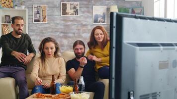 hombre alcanzando para su cerveza mientras acecho televisión con su amigos en el sofá. mujer comiendo Pizza y papas fritas. video