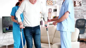 Retired old man in nursing home getting help to walk with crutches from professional social workers video