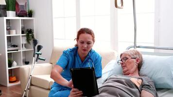 Old woman lying in bed learning from female doctor to use tablet computer. video