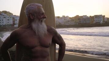 un vecchio uomo con un' barba e in piedi su un' spiaggia con un' tavola da surf a tramonto video