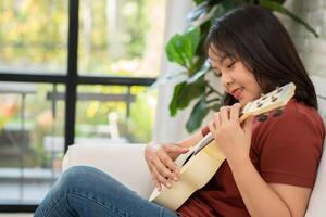 contento asiático mujer sentado en sofá en vivo habitación y practicando ukelele en ocio. relajación y inspiración desde artista. concepto de música, relajarse y pasatiempo actividad disfrutar en ocio foto