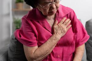 asiático mayor mayor abuela sufrimiento desde corazón dolor y participación su cofre mientras sentado en el sofá en su vivo habitación. médico emergencia tratamiento y salud cuidado seguro proteccion concepto foto