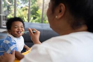 Asian mother feeding her 9 months old her cute little baby and African American helping for holding food plate At Home. Photo series of family, kids and happy people concept. Parents feed kids.