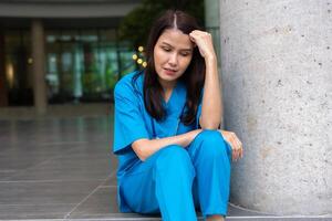 Portrait of stressed and overworked female doctor medical worker surgeon sitting on the floor near the operating room in the hospital and take off the mask photo