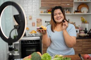 Asian Pregnant blogger looking at smartphone camera, she is live streaming cooking class for pregnant. Asian woman standing happily smiling at the kitchen counter preparing fresh organic salad. photo