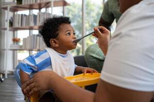 asiático madre alimentación su 9 9 meses antiguo su linda pequeño bebé y africano americano Ayudar para participación comida plato a hogar. foto serie de familia, niños y contento personas concepto. padres alimentar niños.