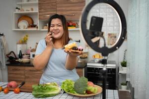 Asian Pregnant blogger looking at smartphone camera, she is live streaming cooking class for pregnant. Asian woman standing happily smiling at the kitchen counter preparing fresh organic salad. photo