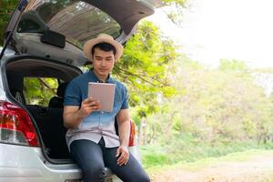 Asian man sitting back of car and holding tables check location and find place to travel. Happy road trip and vacation in summer in the countryside. Concept of journey on leisure, road trip vacation photo