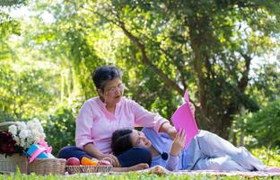 asiático hija dormido en madre regazo y leyendo libro en picnic estera en el parque. un contento mayor mujer negociaciones con su hija. concepto de cuidado de la salud y mayor cuidador apoyo servicio. foto