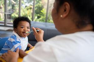 Asian mother feeding her 9 months old her cute little baby and African American helping for holding food plate At Home. Photo series of family, kids and happy people concept. Parents feed kids.