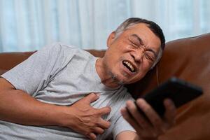 Senior male asian presses hand to his chest has a heart attack suffers from unbearable pain in the living room, lonely men try using the smartphone to call for Emergency help. photo