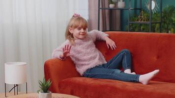 Child girl lying on sofa at home looking at camera, smiling, waving hands gesturing hello or goodbye video