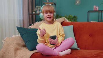 Child girl kid with smartphone in headphones dancing singing listening music at home alone on sofa video