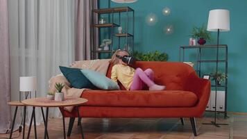 Toddler girl sitting on home sofa using virtual reality headset helmet app to play simulation game video