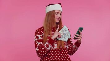 menina dentro vermelho Natal suéter usando Móvel telefone, aguarde dinheiro dólar dinheiro, fazendo conectados presente compras video