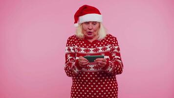 Worried grandmother in Christmas sweater enthusiastically playing racing video games on mobile phone