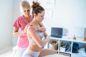 Focused osteopath massaging back of female client sitting on bed in clinic photo