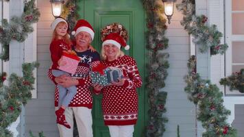 Happy senior grandparents with granddaughter holding many gift boxes near Christmas house, celebrate video