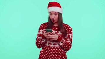 jovem adolescente menina dentro Natal suéter olhando Smartphone exibição Atenciosamente alegria ganhar sucesso sorte video