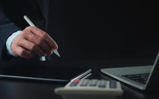 Businessman in black suit working on laptop computer and tablet and calculator, Hand Hands touch on tablet at office with dark background, Online working, Close up, Copy space. photo