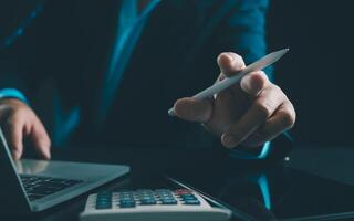 Businessman in black suit working on laptop computer and tablet and calculator, Hand Hands touch on tablet at office with dark background, Online working, Close up, Copy space. photo