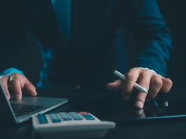 Businessman in black suit working on laptop computer and tablet and calculator, Hand Hands touch on tablet at office with dark background, Online working, Close up, Copy space. photo