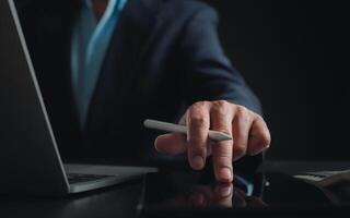 Businessman in black suit working on laptop computer and tablet, Hand Hands touch on tablet at office with dark background, Online working, Close up, Copy space. photo