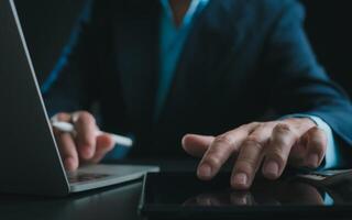 Businessman in black suit working on laptop computer and tablet, Hand Hands touch on tablet at office with dark background, Online working, Close up, Copy space. photo