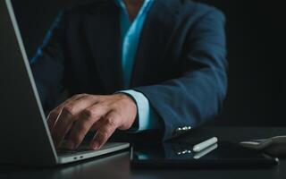 Businessman in black suit working on laptop computer and tablet, Hand typing on keyboard on table at office with dark background, Online working, Close up, Copy space. photo