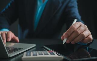 Businessman in black suit working on laptop computer and tablet and calculator, Hand Hands touch on tablet at office with dark background, Online working, Close up, Copy space. photo