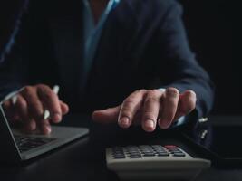 Businessman in black suit working on laptop computer and tablet and calculator, Hand Hands touch on tablet at office with dark background, Online working, Close up, Copy space. photo