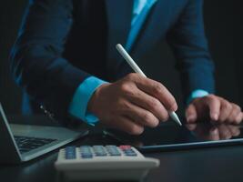 Businessman in black suit working on laptop computer and tablet and calculator, Hand Hands touch on tablet at office with dark background, Online working, Close up, Copy space. photo
