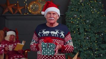 Elderly grandfather in festive sweater presenting Christmas gift box, smiling, looking at camera video