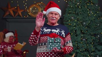 Senior grandfather in Christmas sweater smiling friendly at camera and waving hands gesturing hello video