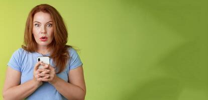 Shocked speechless impressed redhead european middle-aged woman fold lips astonished wow stare camera fascinated react amazed wondered received message hold smartphone intense green background photo