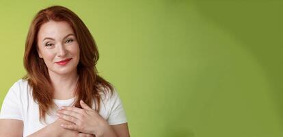 Lovely pleased tender redhead middle-aged ginger woman press hands heart feel touched grateful appreciate sincere heartwarming moment admire thankfully grin look camera glad green background photo