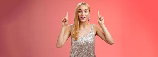 Charming amazed blond european woman in fabulous silver glittering dress raise hands point up amused enjoying watching shooting stars, fireworks gaze camera excited happy surprised, red background photo