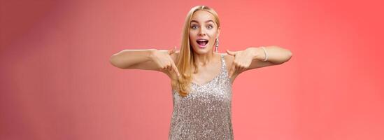 Impressed excited attractive glamour blond girl in silver glittering dress gasping thrilled pointing down glance camera fascinated check out fabulous awesome promo, standing amused red background photo