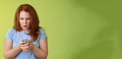 Confused unsure redhead middle-aged woman learn how use social media trying understand emoji look intense focused smartphone display reading important news message stand green background photo