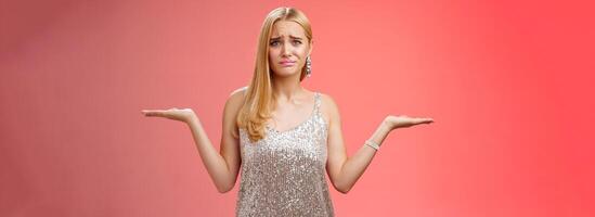 Nervous unsure doubtful cute blond woman struggle make decision shrugging pointing sideways frowning upset standing insecure feel pressure cannot decide choice make, frustrated red background photo