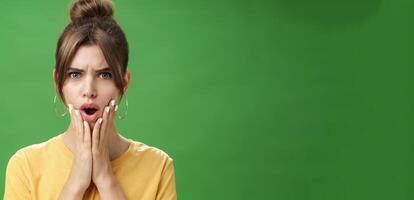 Shocked woman reacting to pimple looking disturbed and displeased in mirror holding hands pressed to cheeks open mouth and frowning being disappointed and upset over green background photo