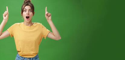 Waist-up shot of interested and surprised amazed good-looking female in yellow t-shirt with tattoo dropping jaw impressed and astonished raising hands pointing up questioned against green background photo