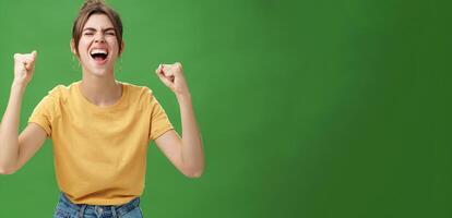 Waist-up shot of charismatic energized and excited female in yellow t-shirt closing eyes yelling from joy and happiness raising hands in cheer celebrating successful news over green background photo