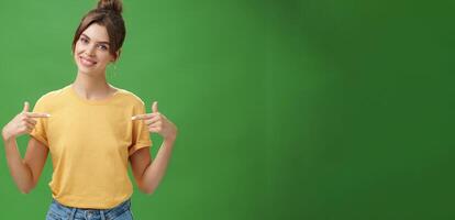 Calm and polite friendly-looking skillful young female coworker in yellow t-shirt pointing at herself self-assured tilting head and smiling at camera wanting participate in event over green background photo