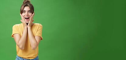 Indoor shot of excited and surprised energized attractive glamorous female in yellow t-shirt touching cheeks from amazement smiling broadly astonished posing over green background photo