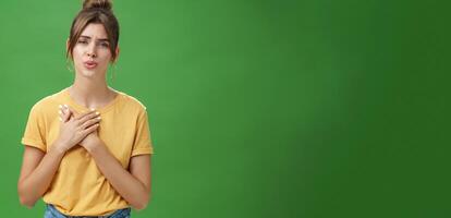 Touched and pleased attractive young woman in yellow t-shirt holding palms pressed to chest in thankful and grateful gesture folding lips melting heart seeing cute thing over green background photo