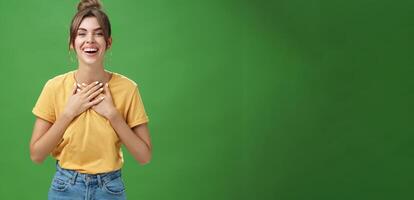 Delighted charming woman with gapped teeth in cozy outfit holding palms on chest in grateful or thankful pose smiling broadly receiving warm congratulations standing satisfied, happy over green wall photo