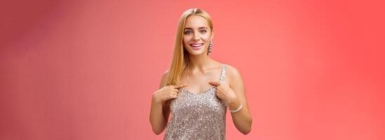 Boastful good-looking confident blond european woman in silver luxurious dress pointing herself smiling proudly bragging talking own accomplishments goals standing red background self-assured photo